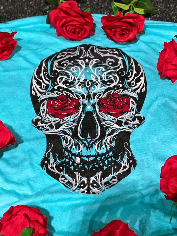 Turquoise shirt with sugar skull on it with roses laying around the shirt. 