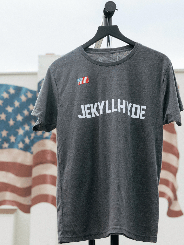 Grey shirt with Jekyllhyde text on a hanger with the american flag in the background. 