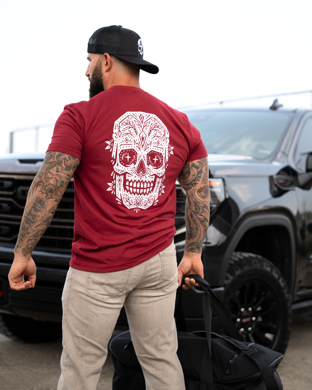 A man wearing a maroon t-shirt with a large white sugar skull graphic on the back, standing in front of a black truck and pulling a black duffel bag.