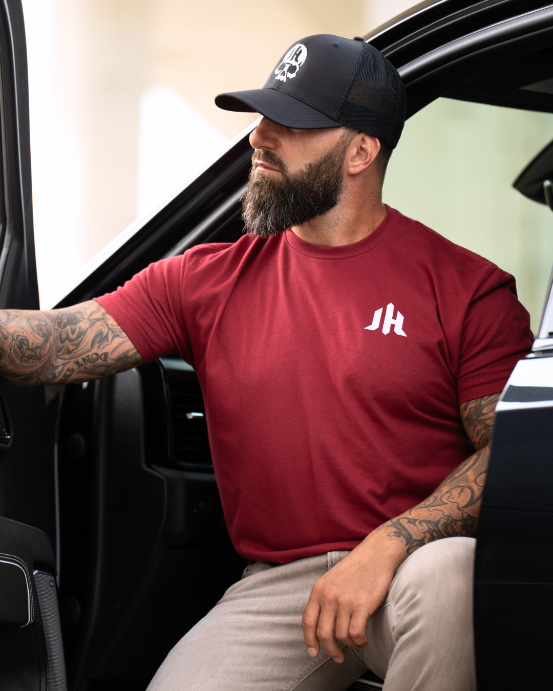 A man with a beard and tattoos sitting in a vehicle doorway, wearing a maroon short-sleeve t-shirt with a white stylized 'JH' logo on the left chest, a black baseball cap, and khaki pants.