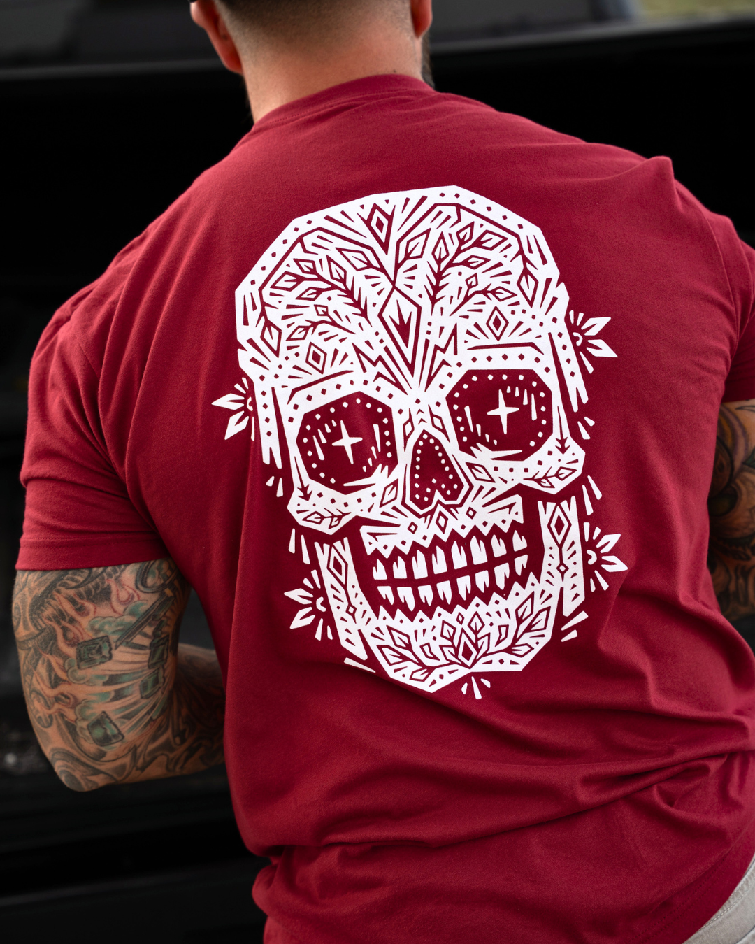 A man viewed from behind wearing a red t-shirt with a large white sugar skull graphic on the back, set against a dark outdoor background.