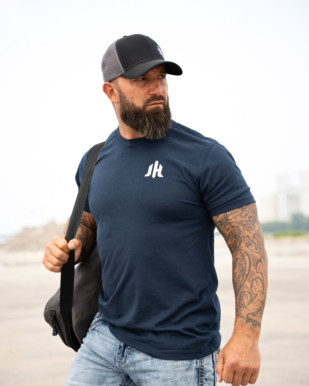 A man wearing a navy blue crewneck t-shirt with a white stylized 'JH' logo on the chest, a black and gray trucker hat, and light blue jeans, carrying a black shoulder bag on a beach.