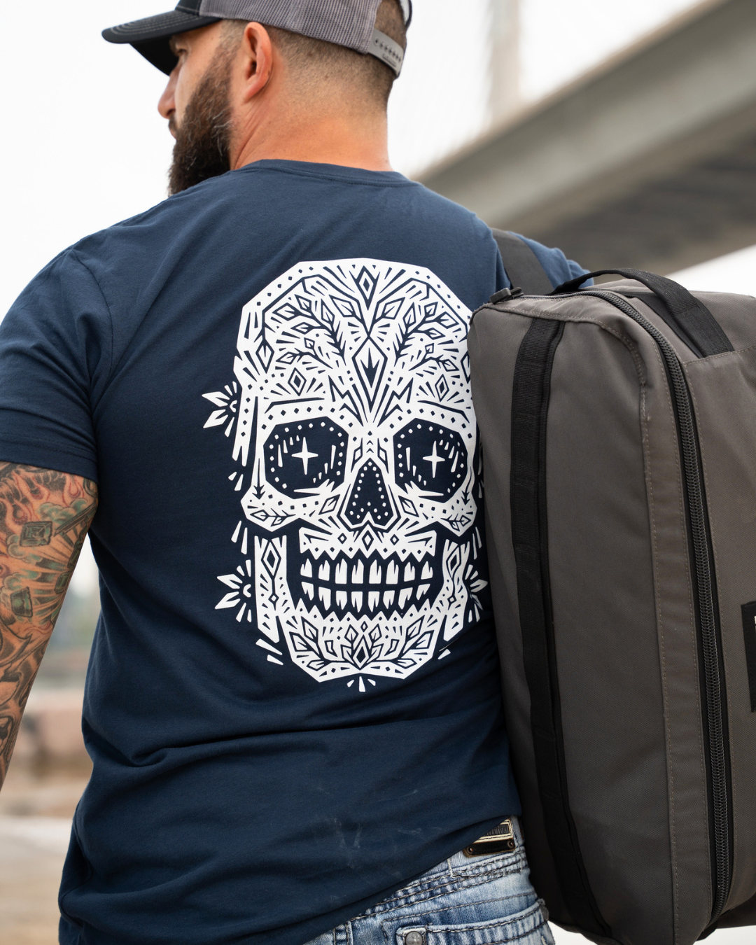 A man viewed from behind wearing a navy blue t-shirt with a large white sugar skull graphic on the back, carrying a dark gray backpack.