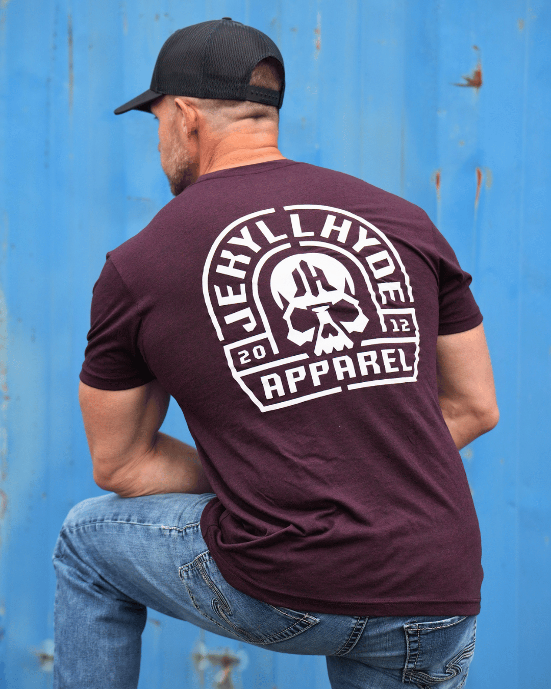 muscular man standing in front of a blue background with a maroon shirt with skull on it. 