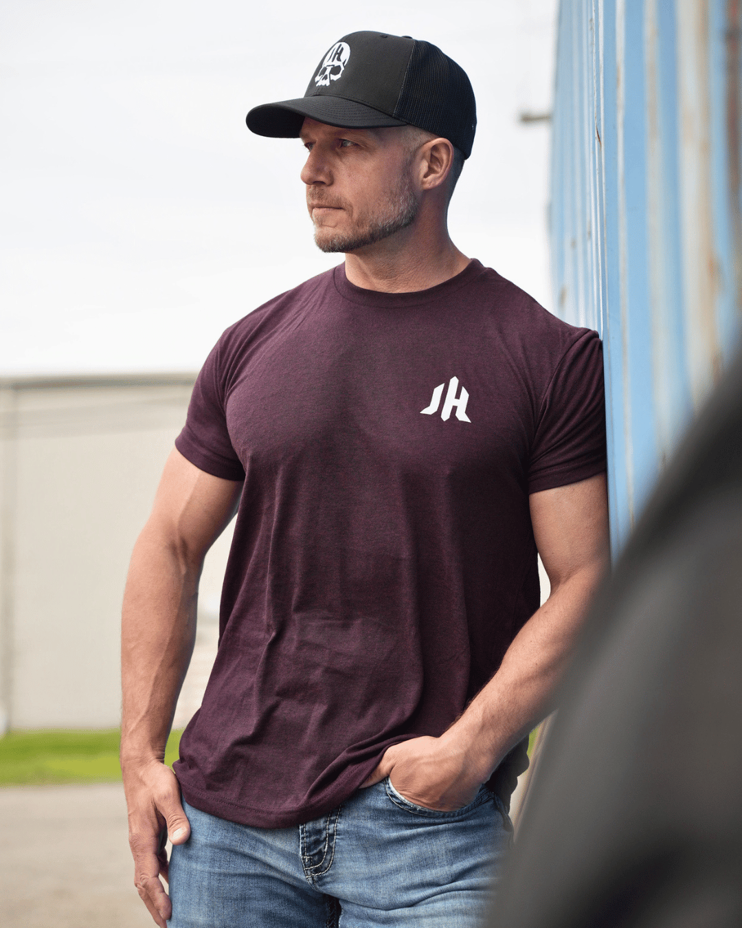 Man leaning against a wall with wearing a maroon tshirt with JH on the left chest. 