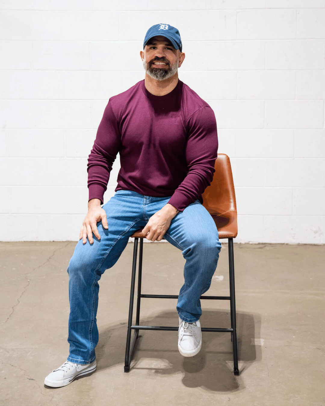 A bearded man sitting on the edge of a chair wearing a maroon thermal. 