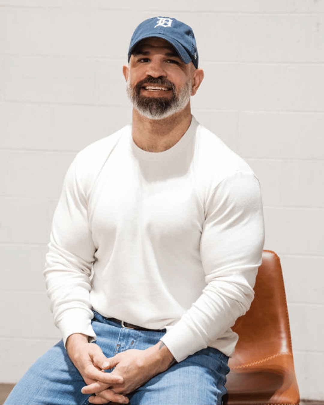 A man sitting in a chair wearing an off white thermal and a blue hat. 