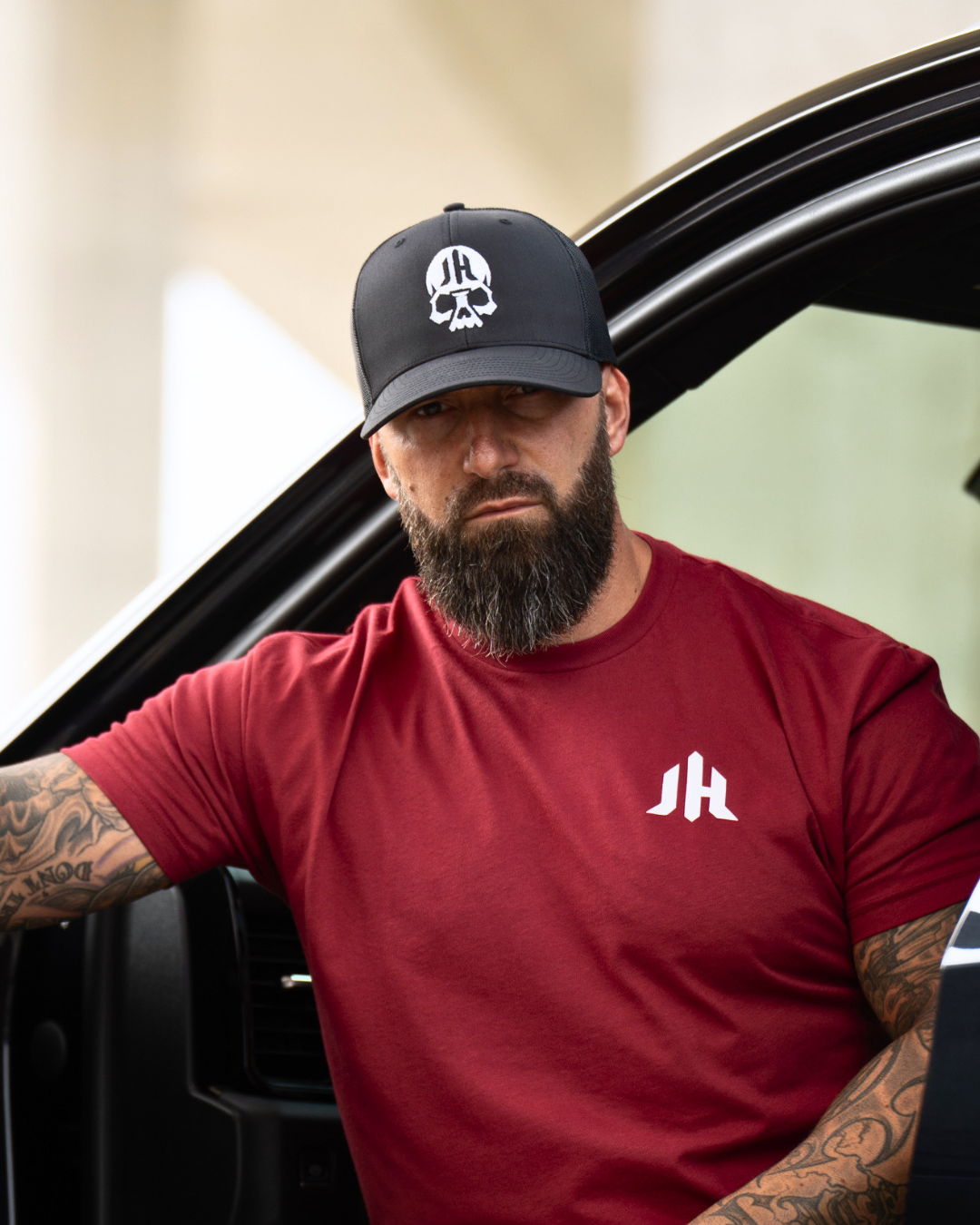 A man with a beard and tattoos leans against a vehicle door wearing a black baseball cap with a white skull logo and a red t-shirt with a white 'JH' logo.