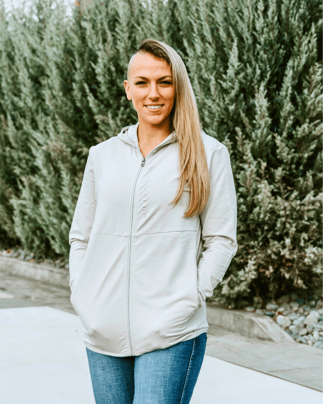 Woman standing in a tan jacket with a tree lined background. 