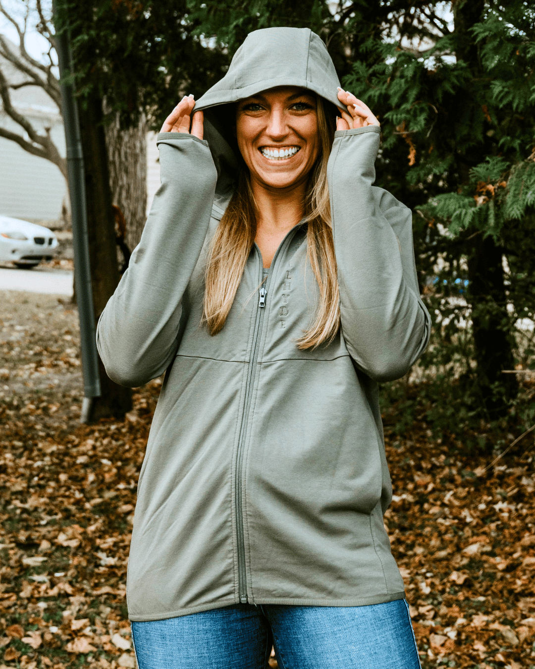 Woman standing in front of trees wearing a green jacket with the hood pulled up. 