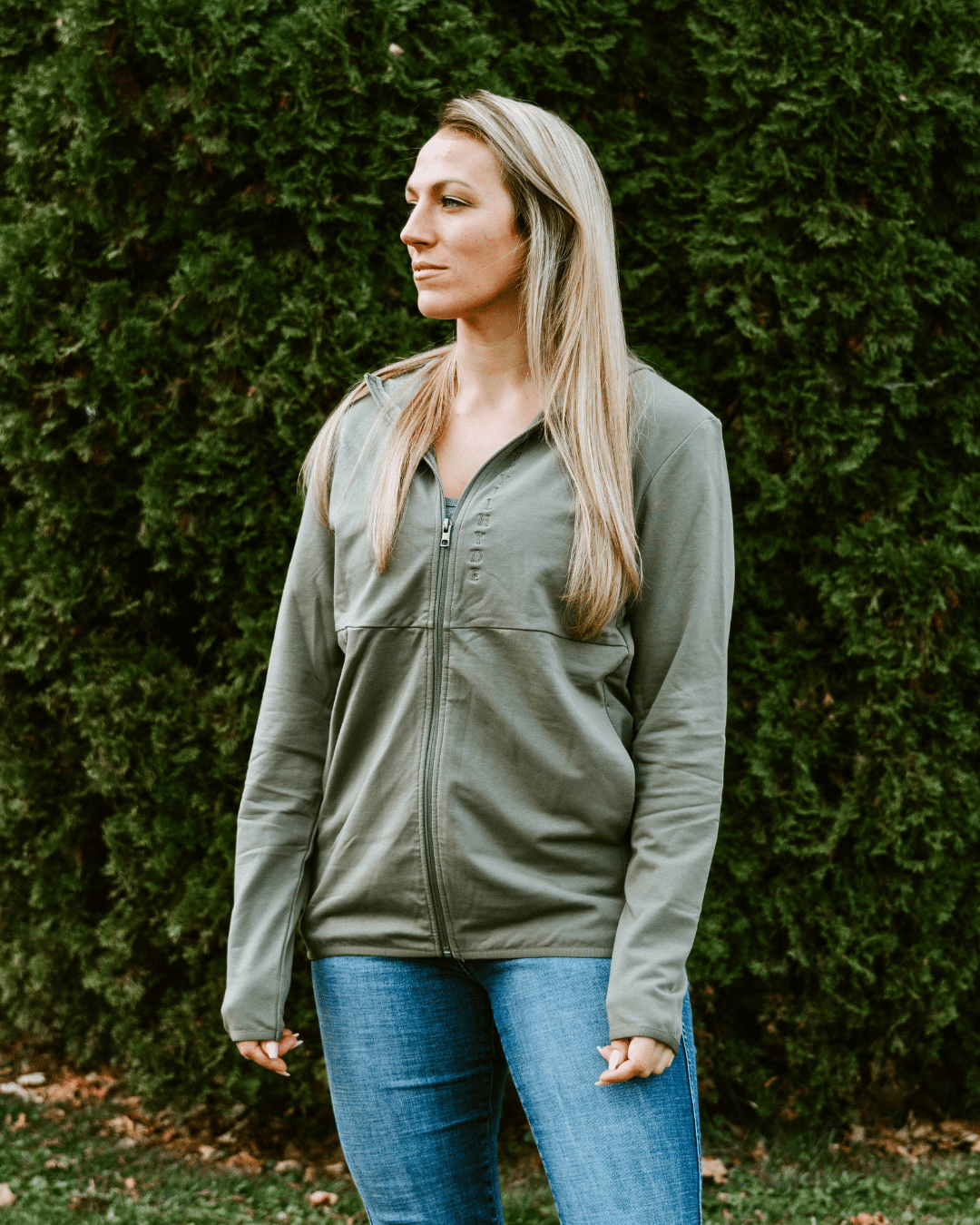 Woman standing in a green jacket with a tree lined background. 