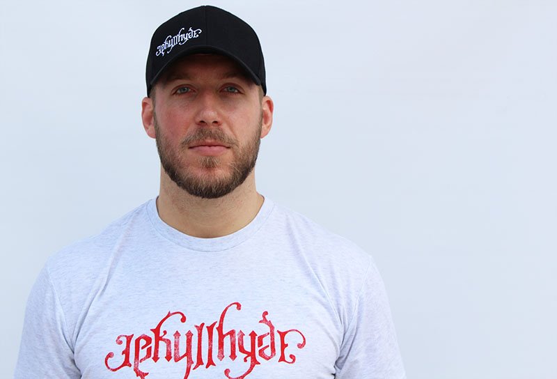 Man looking into the camera wearing a black baseball cap with a jekyllhyde logo in white. 