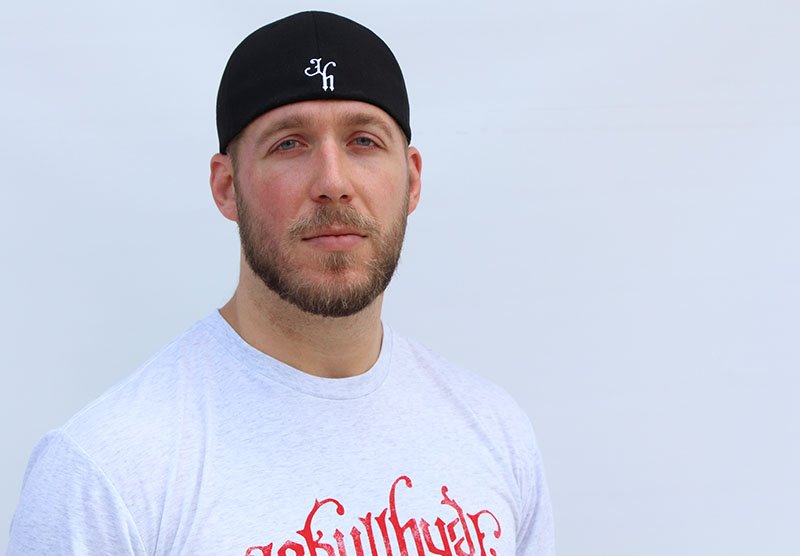 Man looking into the camera wearing a backwards black baseball cap. 