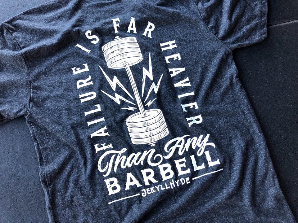 Black shirt on the floor with a barbell in white screen print. 