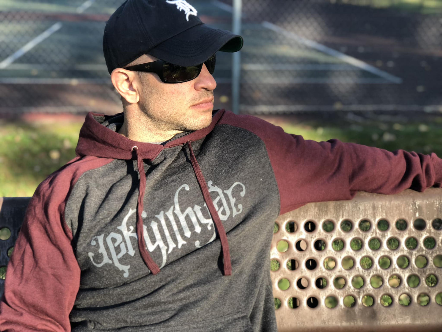man sitting on a park bench in a maroon and charcoal hoodie, wearing sunglasses. 