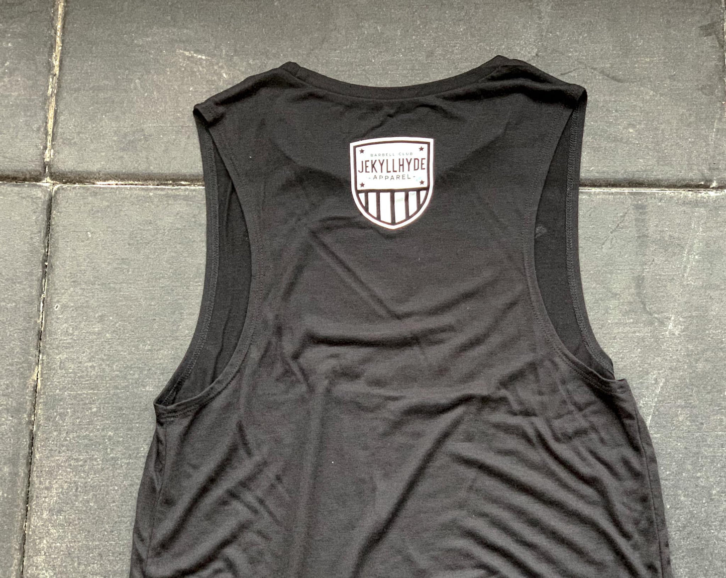 A black muscle tee laying on the ground with a white jekyllhyde shield. 