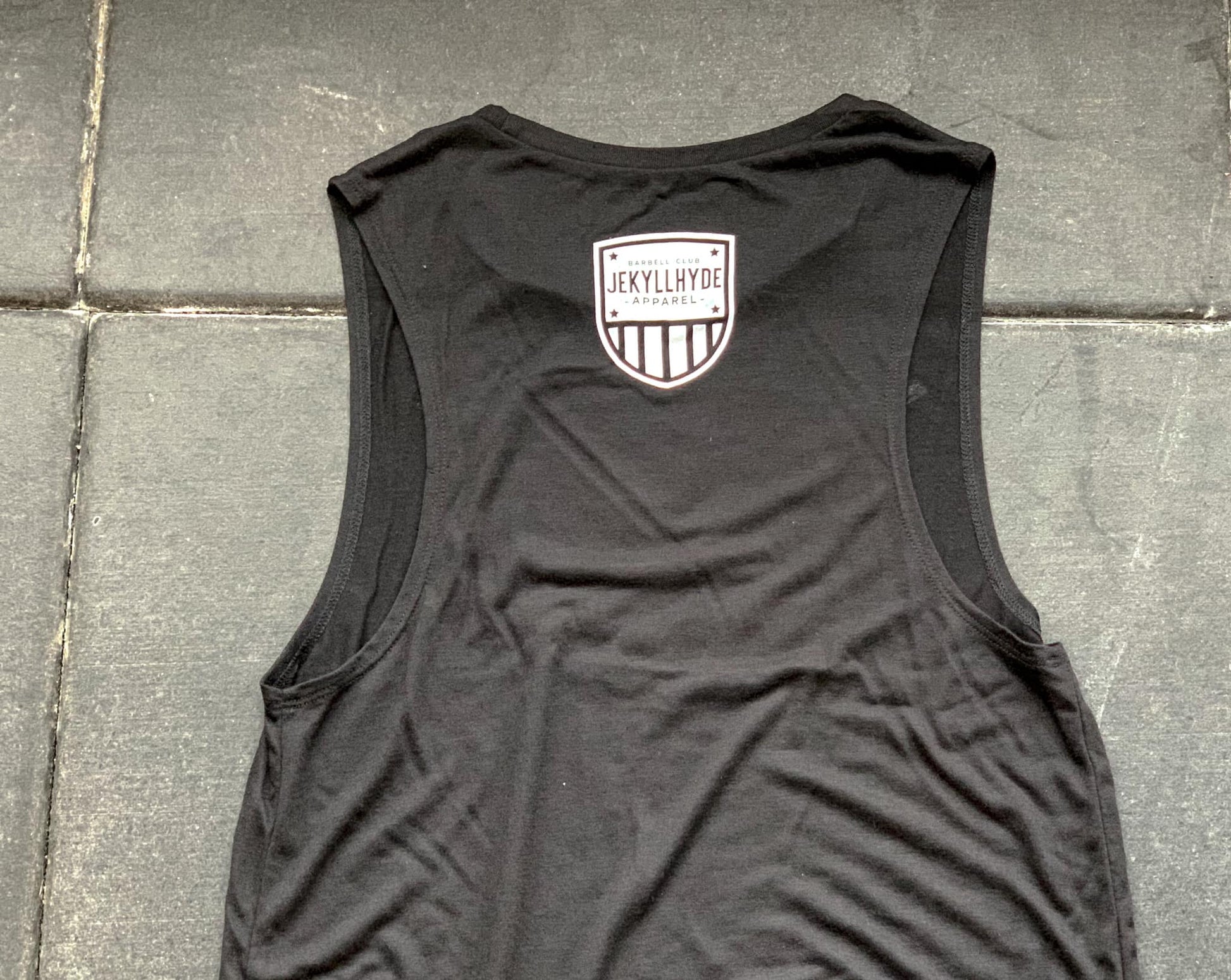 A black muscle tee laying on the ground with a white jekyllhyde shield. 
