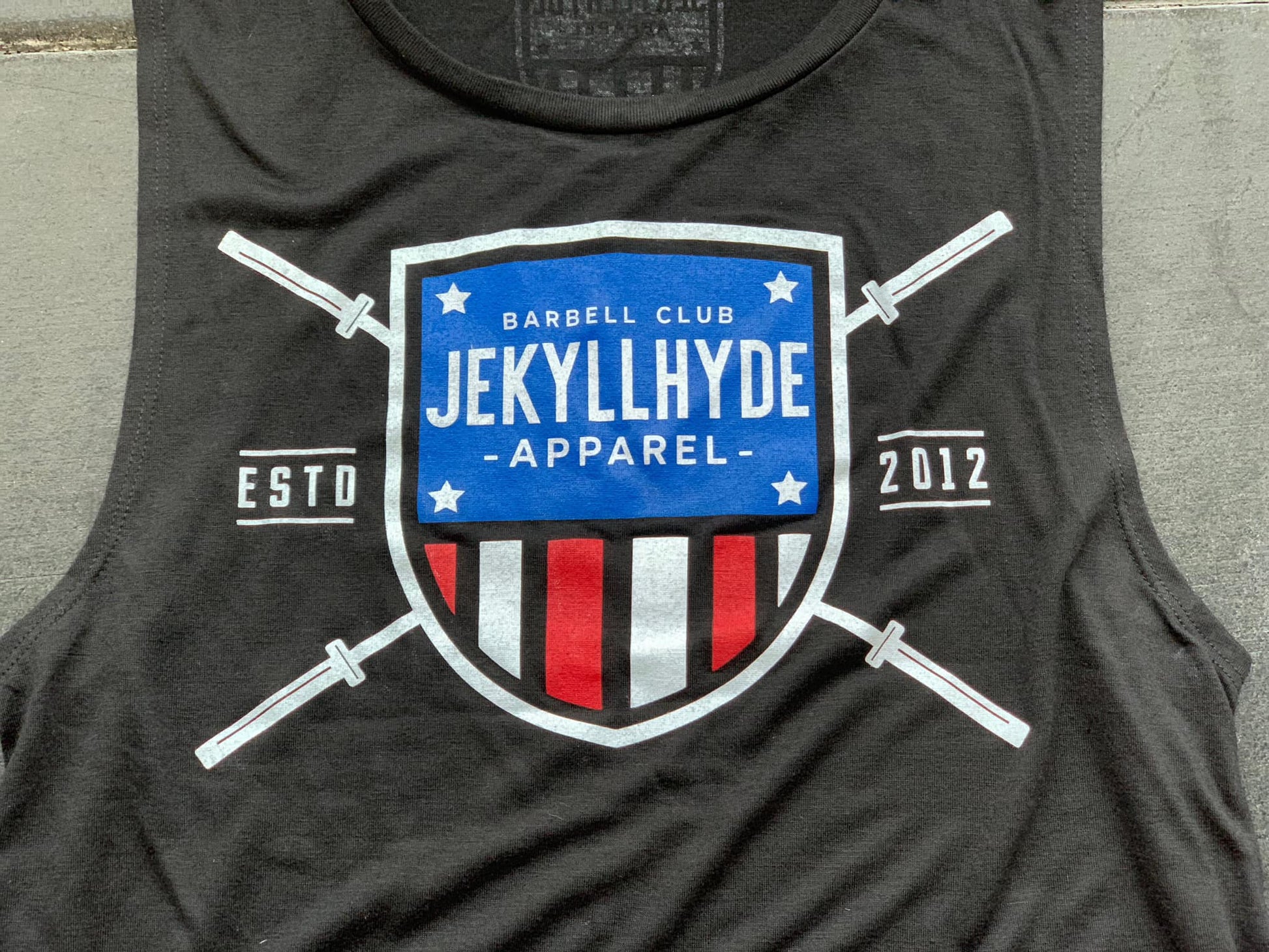 A black muscle tee laying on the ground with a barbell shield on it. 
