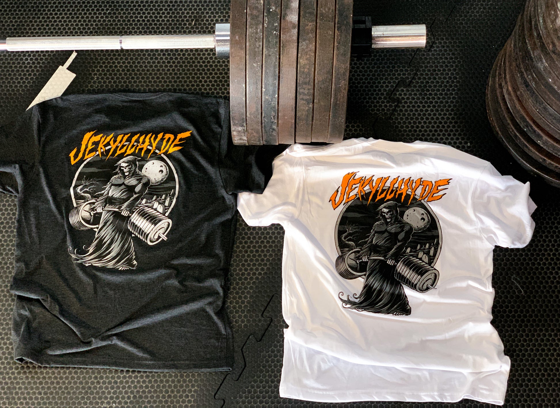 Black and white shirts laying on the ground next to a loaded barbell with a deadlifting grim reaper. 