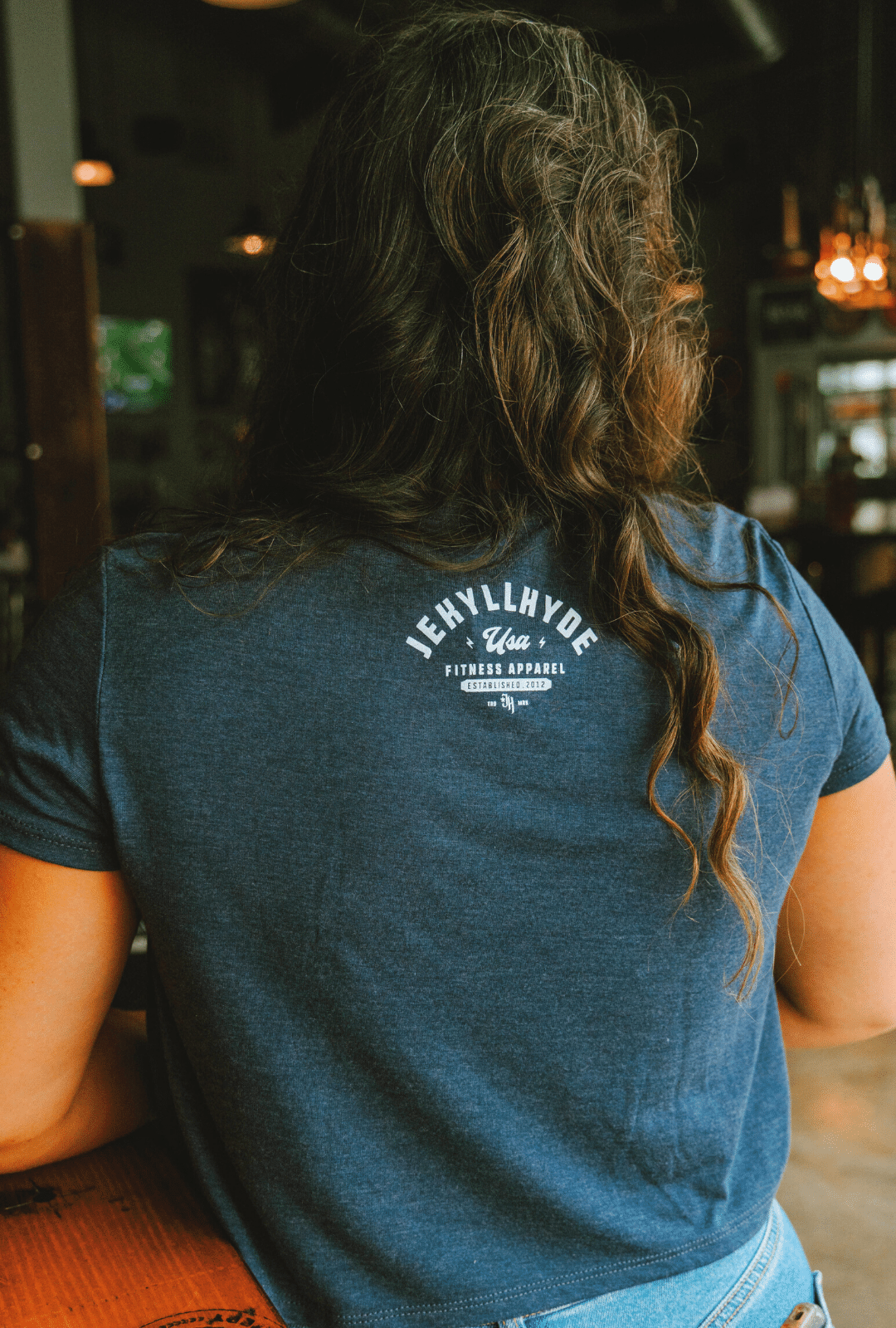 Woman standing at a bar with her back to the camera wearing a blue crop with a Jekyllhyde logo at the top of the neck. 