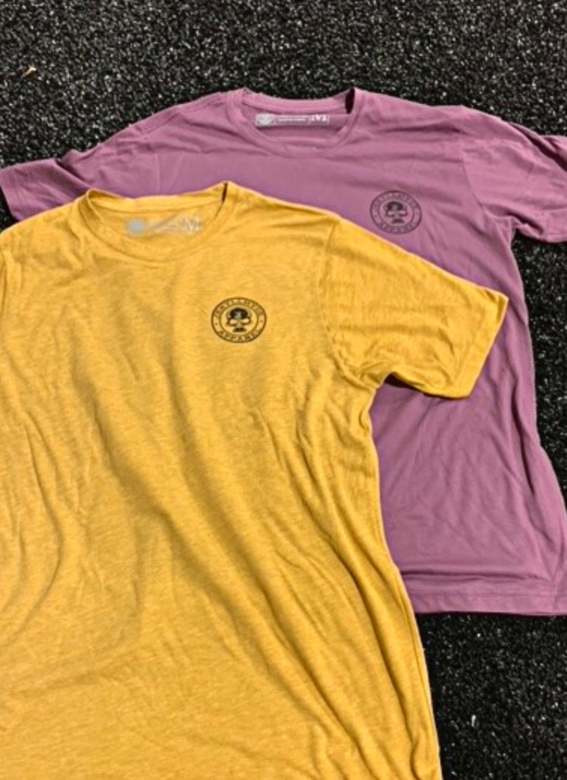 A purple and gold tshirt with a jekyllhyde logo on the left chest, laying on black turf. 