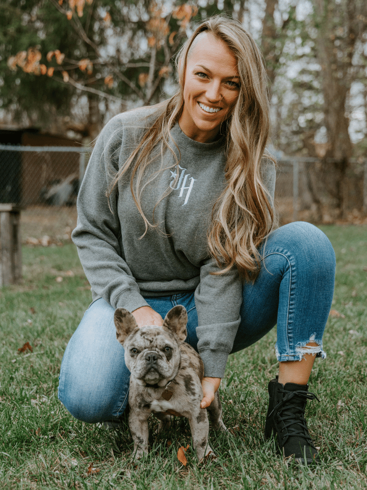 Woman kneeling holding a puppy looking into the camera wearing a grey crewneck sweatshirt. 