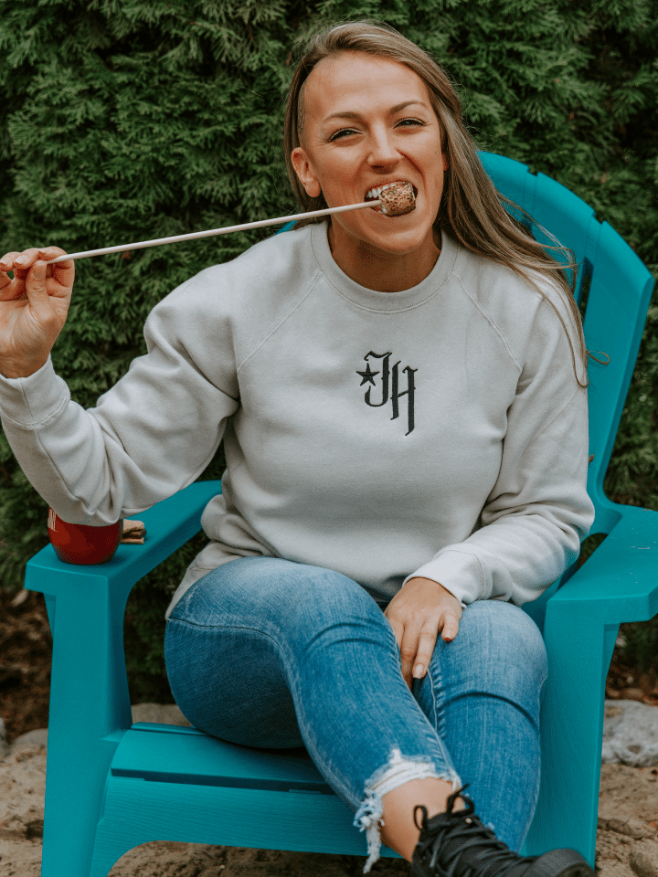 Woman eating a marshmallow off a stick sitting in a chair wearing a sand colored crewneck sweatshirt. 