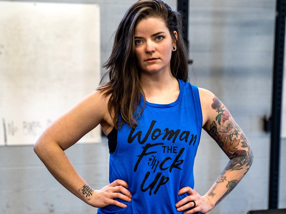 A woman in a blue muscle tee with the words "woman the F*ck up" printed on it. 