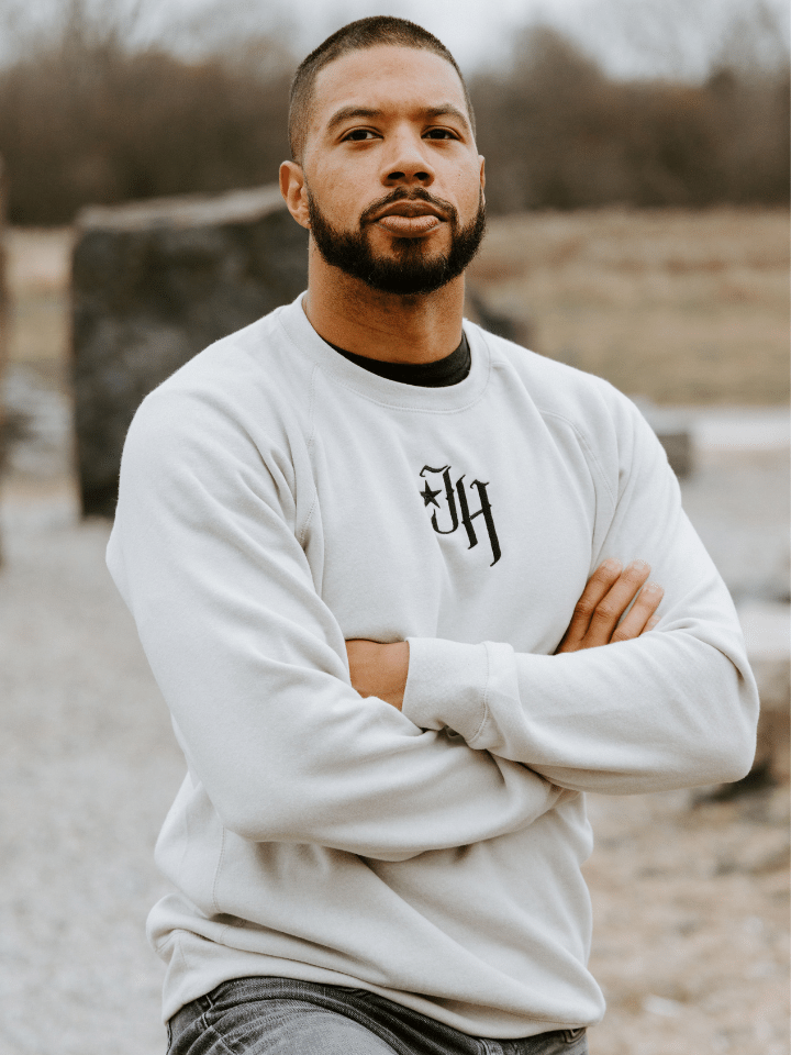 Man standing with his arms crossed looking into the camera wearing a sand colored crewneck sweatshirt. 