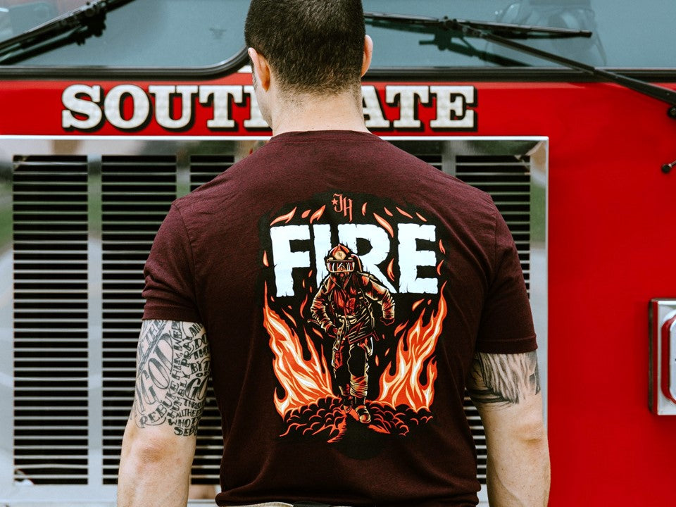 Man standing at a fire truck with maroon tshirt that has a fireman on it. 
