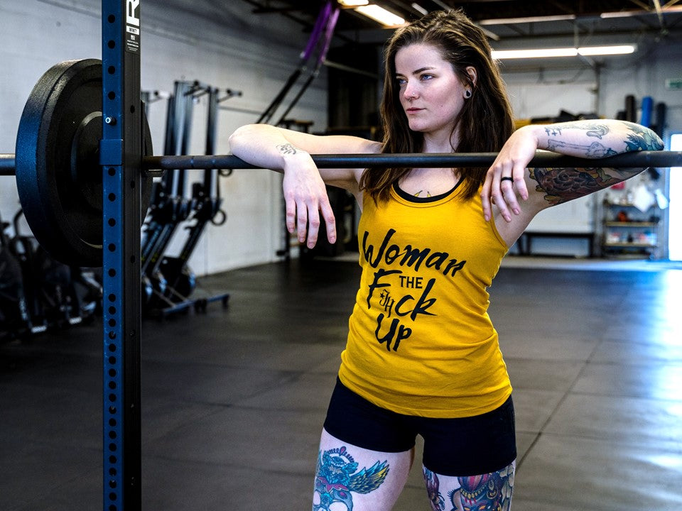 A woman standing at a barbell wearing a gold tank top. 