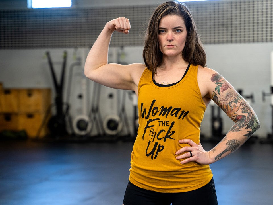 A woman wearing a gold tank top flexing her bicep. 