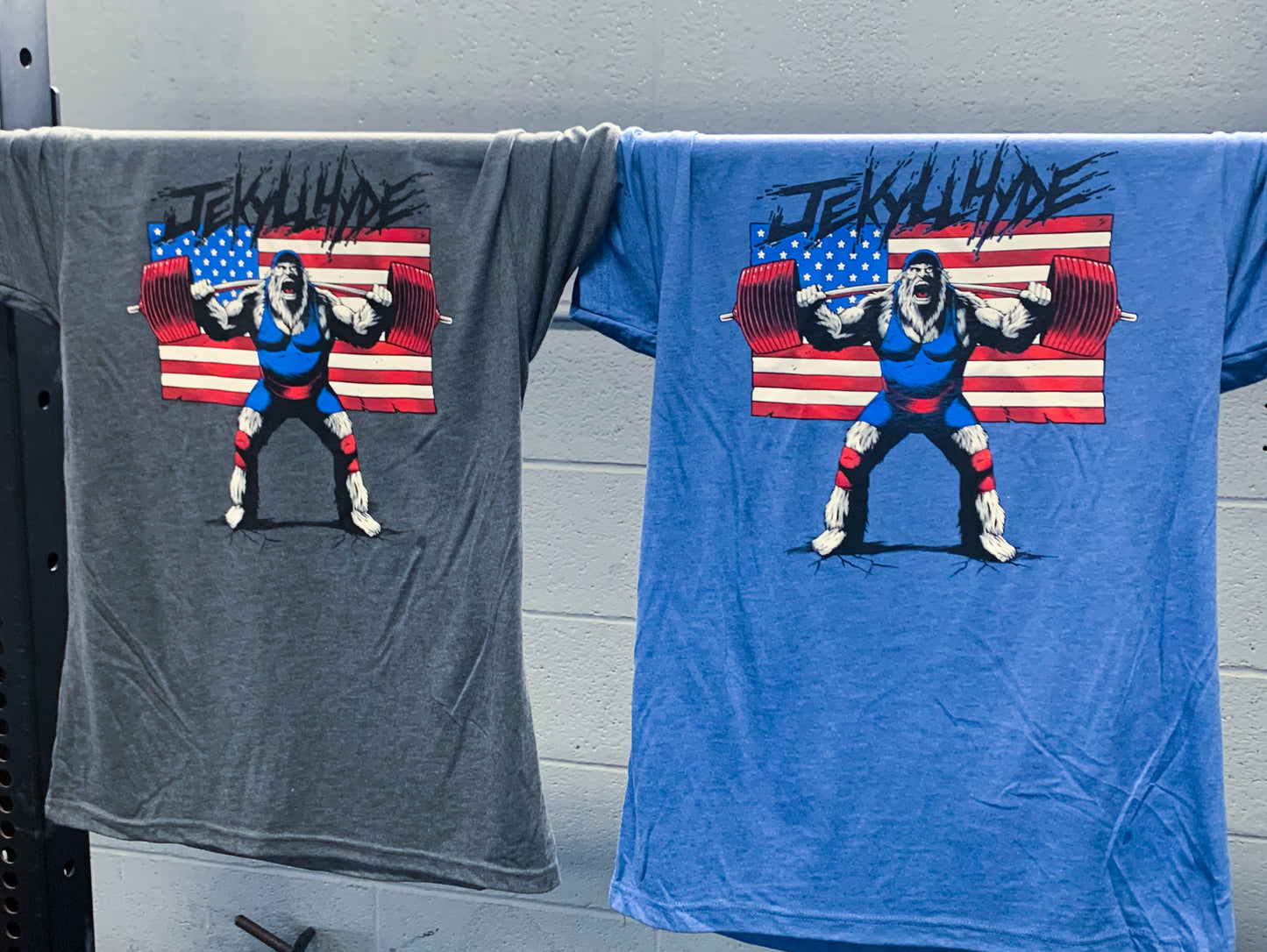 Grey and blue tshirts with a squatting big foot hanging from a barbell. 