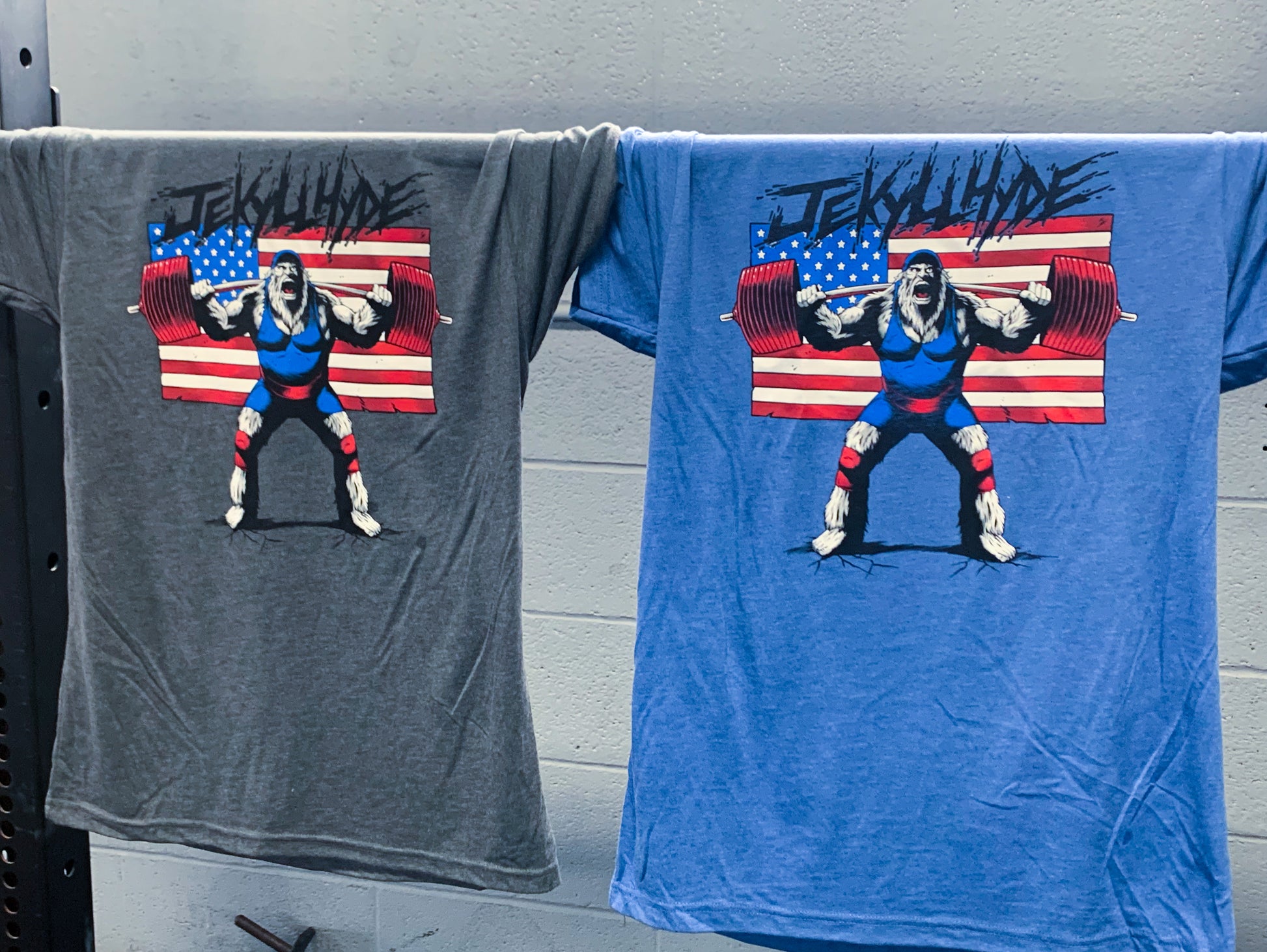 Grey and blue tshirts with a squatting big foot hanging from a barbell. 
