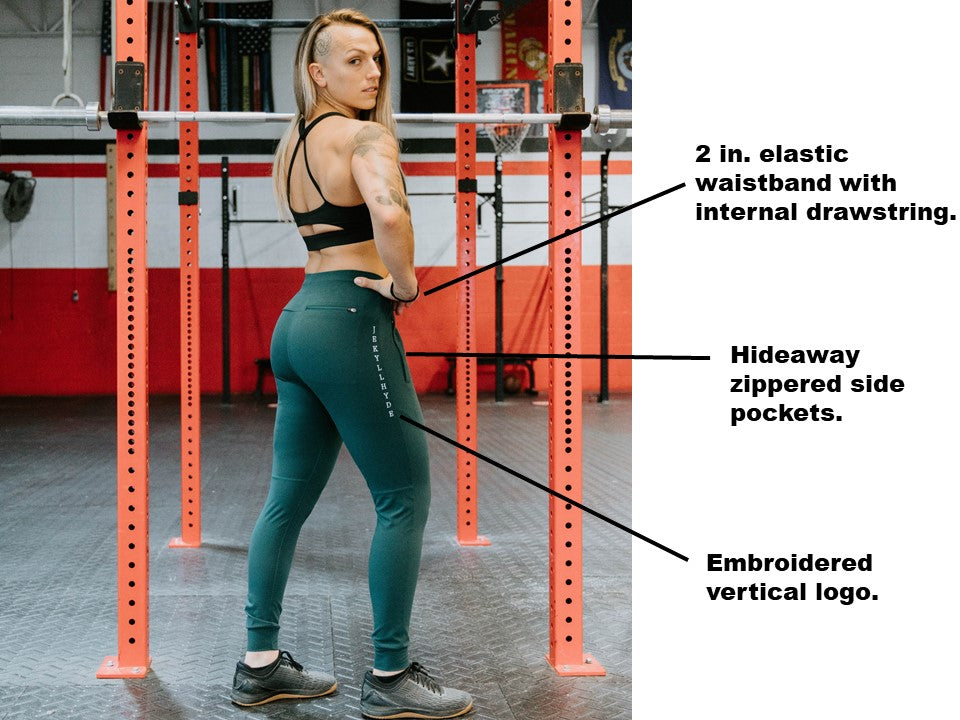 Muscular woman standing at a fitness rig in green joggers with a side bar of text that reads "2 in. elastic waistband with drawstring. Hideaway zippered side pockets, embroidered vertical logo. 