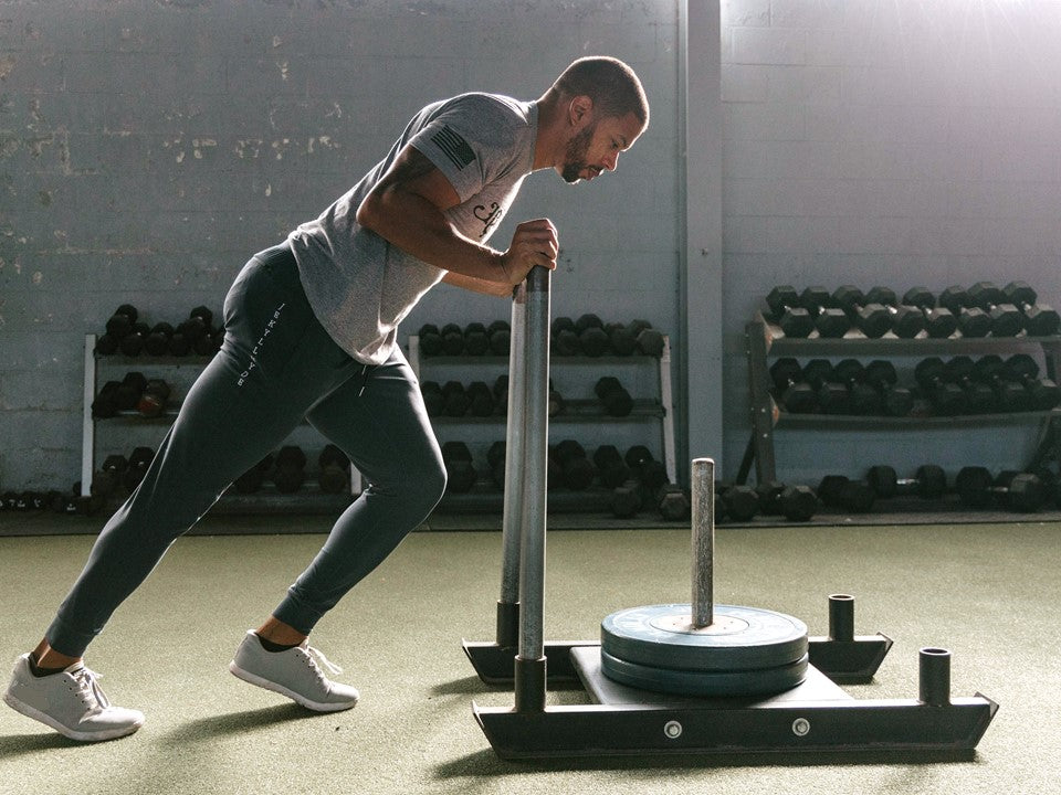 A man pushing a weighted sled on turf in blue joggers.
