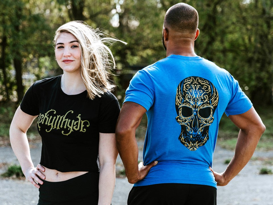 Woman in a cropped tshirt looking forward with Jekyllhyde logo on it. And a man looking away with blue sugar skull tshirt. 