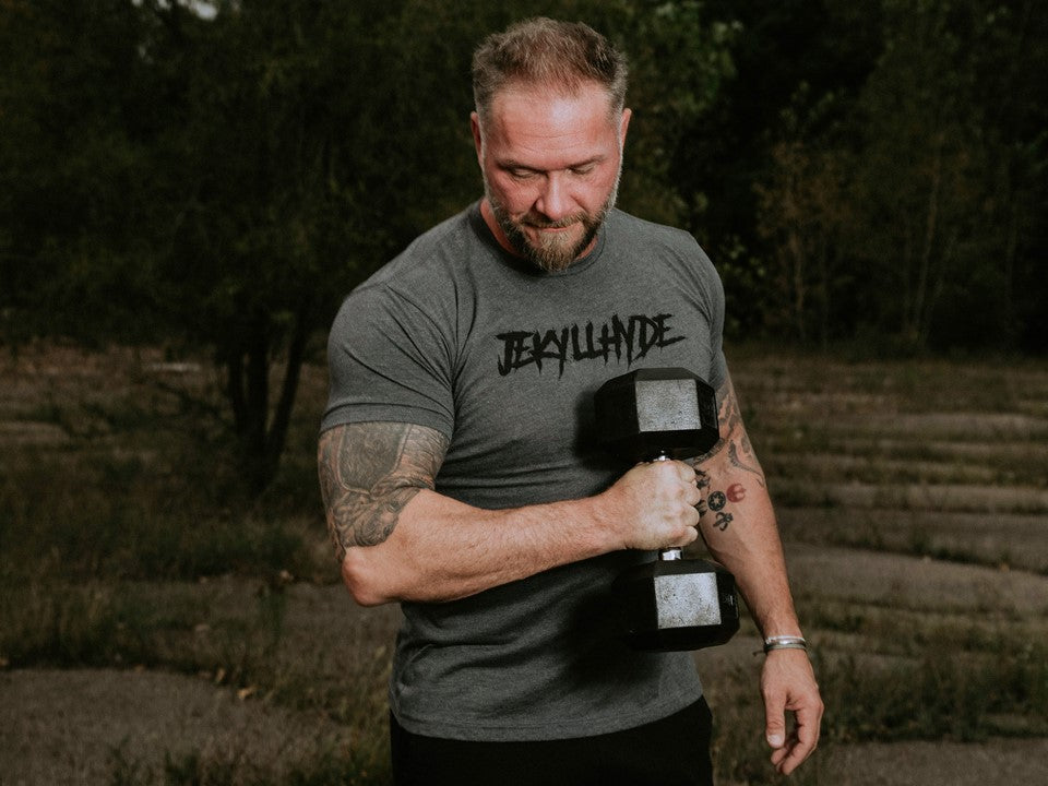 Man in grey shirt curling a dumbbell with Jekyllhyde written across the chest. 