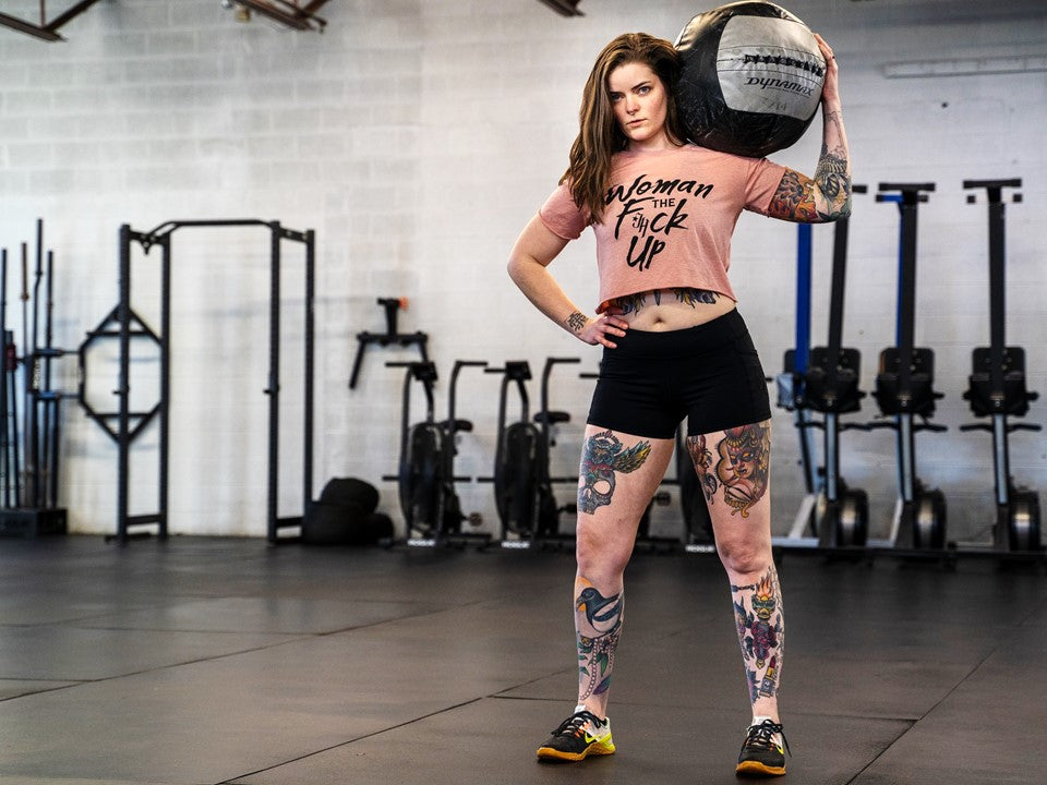 A muscular, tattooed woman holding a medicine ball on her shoulder wearing a pink crop top. 