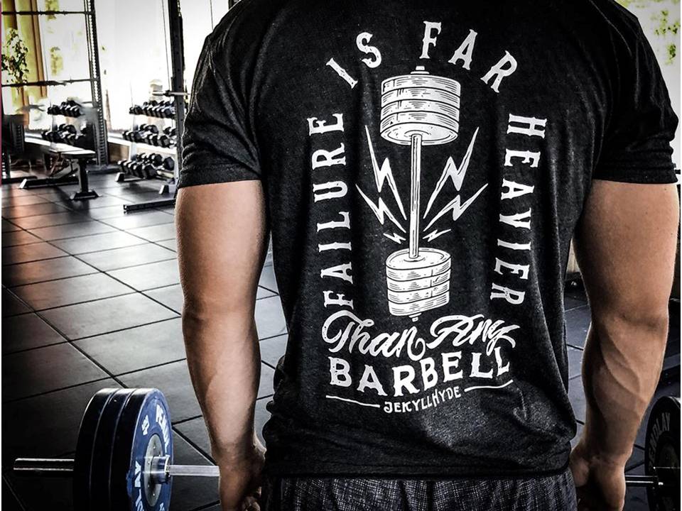 Man standing at a barbell with a black shirt with white text and graphic. 