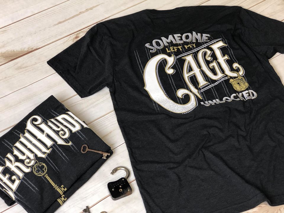 Flat lay of black tshirt with white and brass text that reads; Someone left my cage unlocked. 