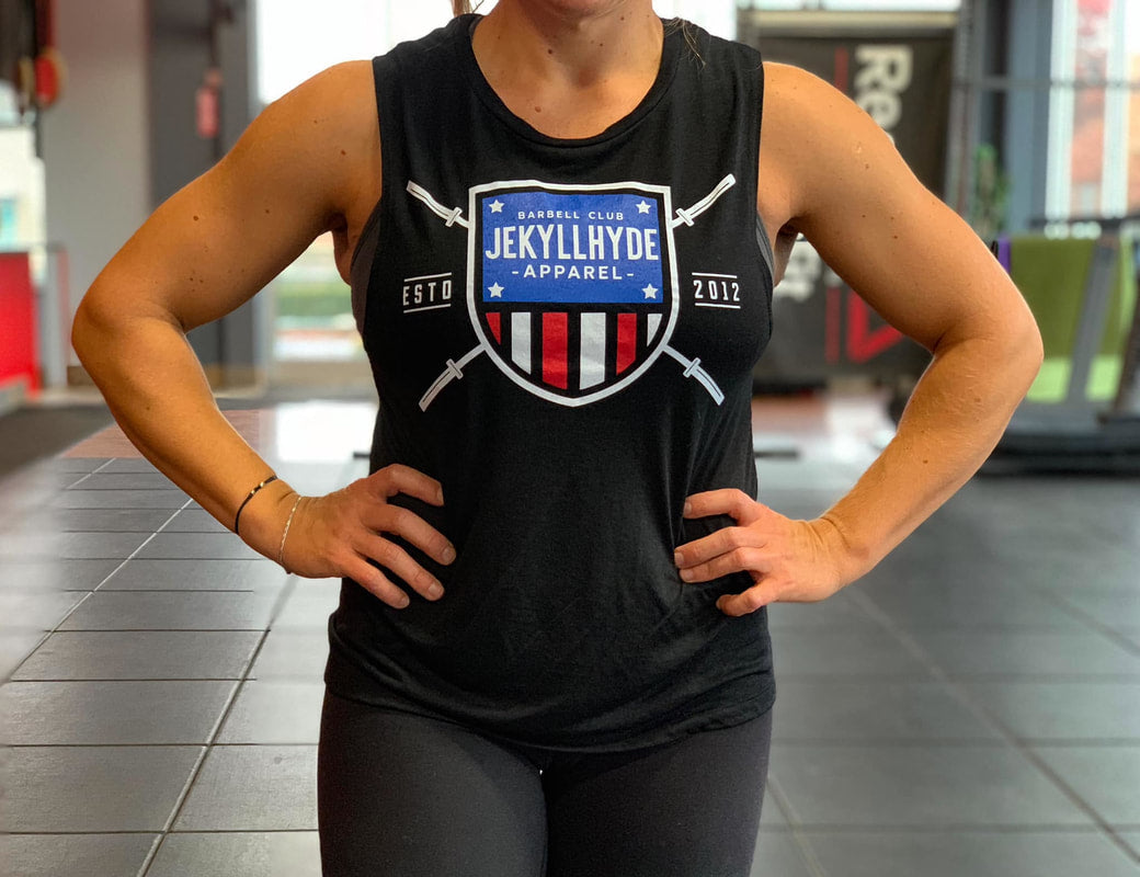 A woman's torso wearing a black muscle tee with a barbell shield on it. 
