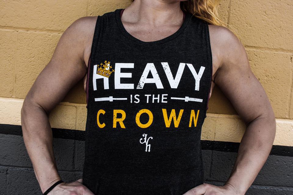 Close us of a black muscle tee with the words "Heavy is the crown" against a brick wall. 