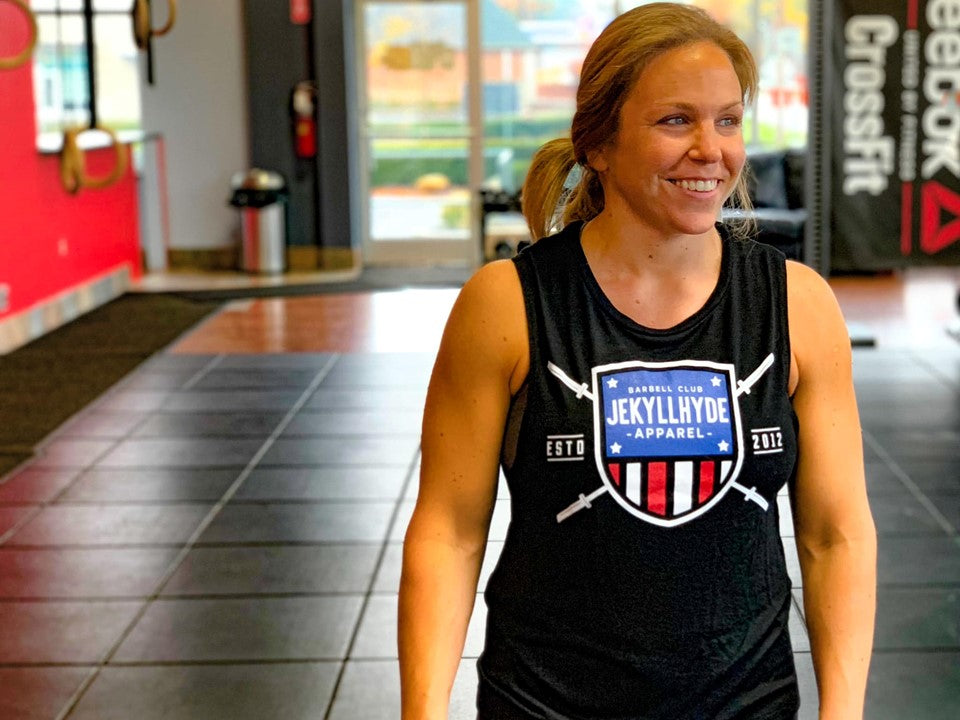 A muscular smiling woman wearing a black muscle tee with a barbell shield. 