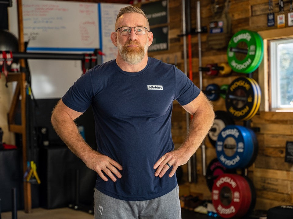 Man standing in a garage gym looking into the camera wearing a blue shirt with a Jekyllhyde logo on the left chest. 