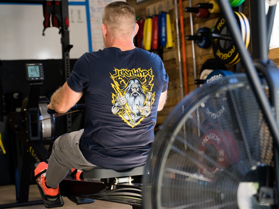 A man on a rowing machine wearing a Jekyllhyde tshirt with Zeus on it. 